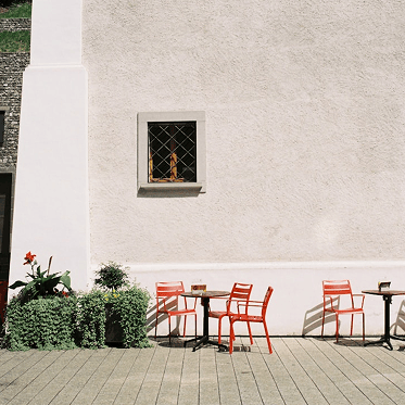 Leere Terrasse mit roten Stühlen und Tischen vor weißer Wand und grünem Pflanzenarrangement.