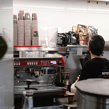 Barista steht vor einer Espressomaschine mit Bechern und Kaffeepackungen auf einem Regal.