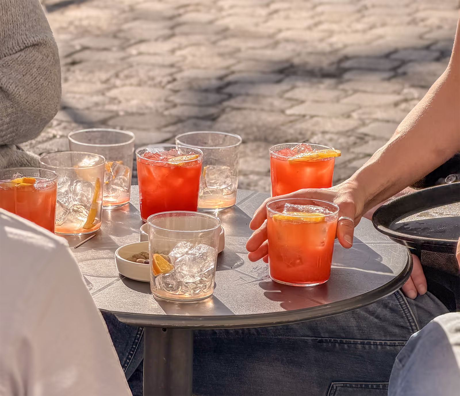 Runder Tisch mit mehreren Aperitivo-Gläsern auf sonniger Terrasse des FINO Freiburg, Hand greift nach Glas