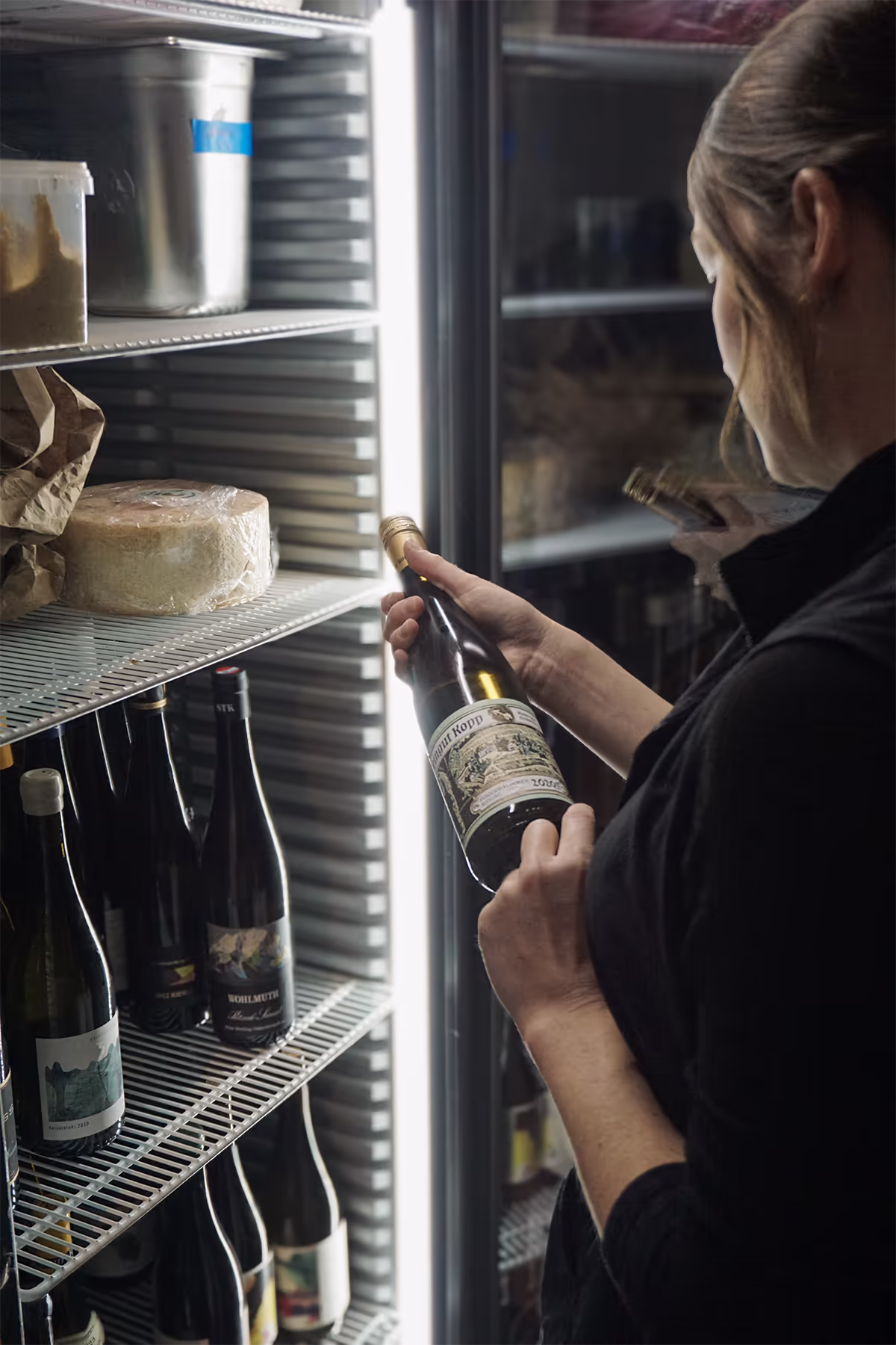 Große Weinauswahl im FINO Freiburg: Frau hält Weinflasche vor gefülltem Kühlschrank mit mehreren Weinflaschen und Käse