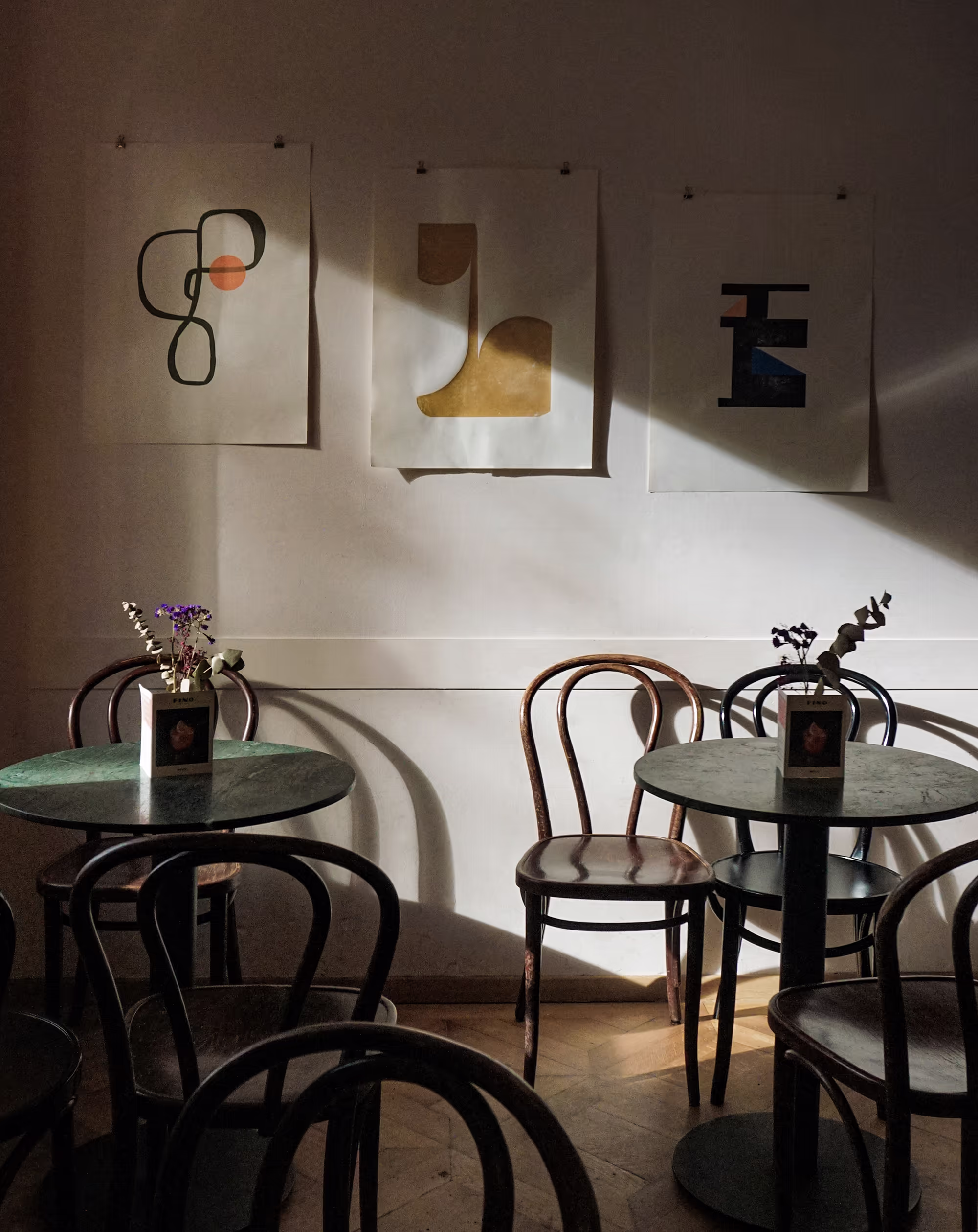 Zwei runde Bistrotische mit Holzstühlen im Innenbereich des Café FINO Freiburg, drei abstrakte Kunstdrucke an der Wand