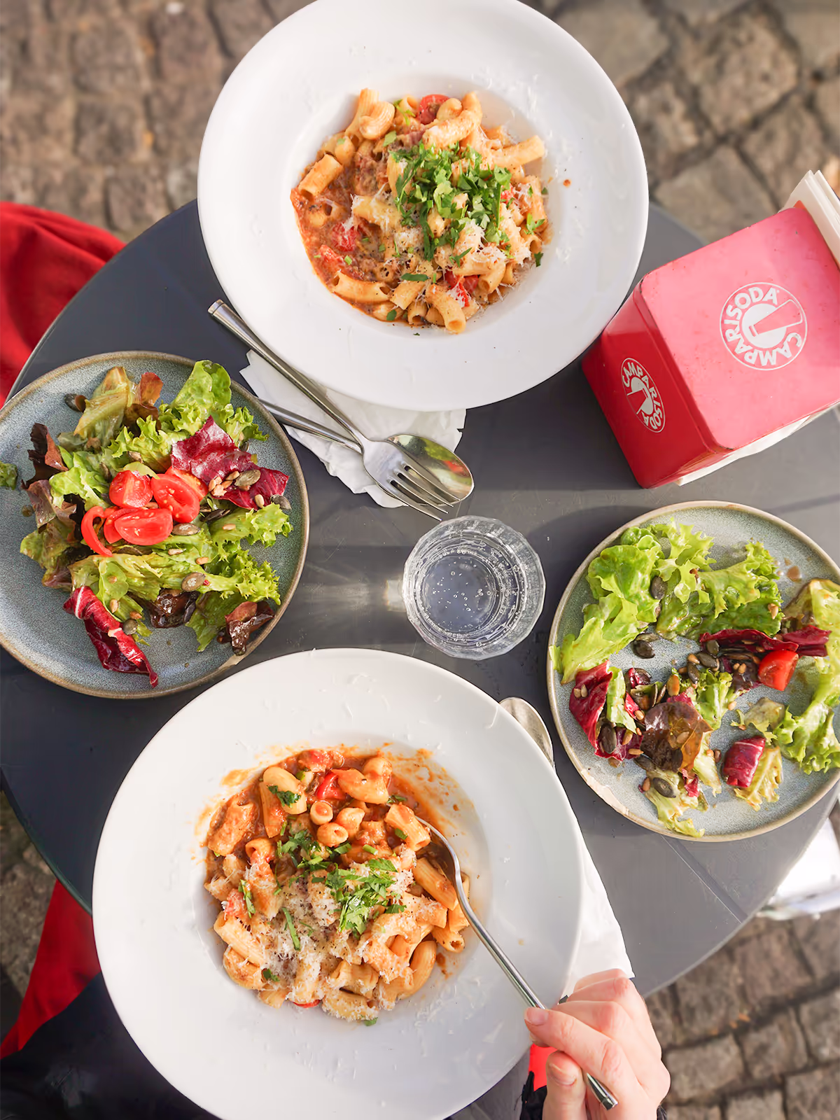 Zwei Teller mit hausgemachter Pasta und zwei Teller mit gemischtem Salat auf einem runden Tisch im Außenbereich vom Café FINO