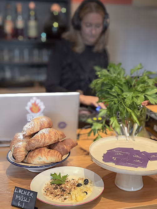 Teller mit Müsli und Croissants auf Tisch, im Hintergrund Person mit Kopfhörern, grüne Pflanzen.