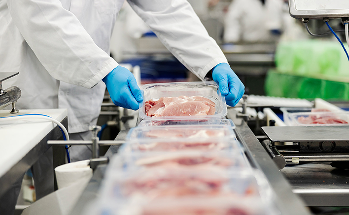 Worker wearing blue gloves and white coat handling packaged raw meat on a conveyor belt in a food processing facility.