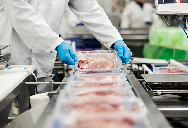 Worker wearing blue gloves and white coat handling packaged raw meat on a conveyor belt in a food processing facility.