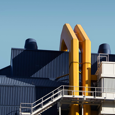 Industrial building facade with large yellow vertical ducts and a metal staircase against a clear blue sky.