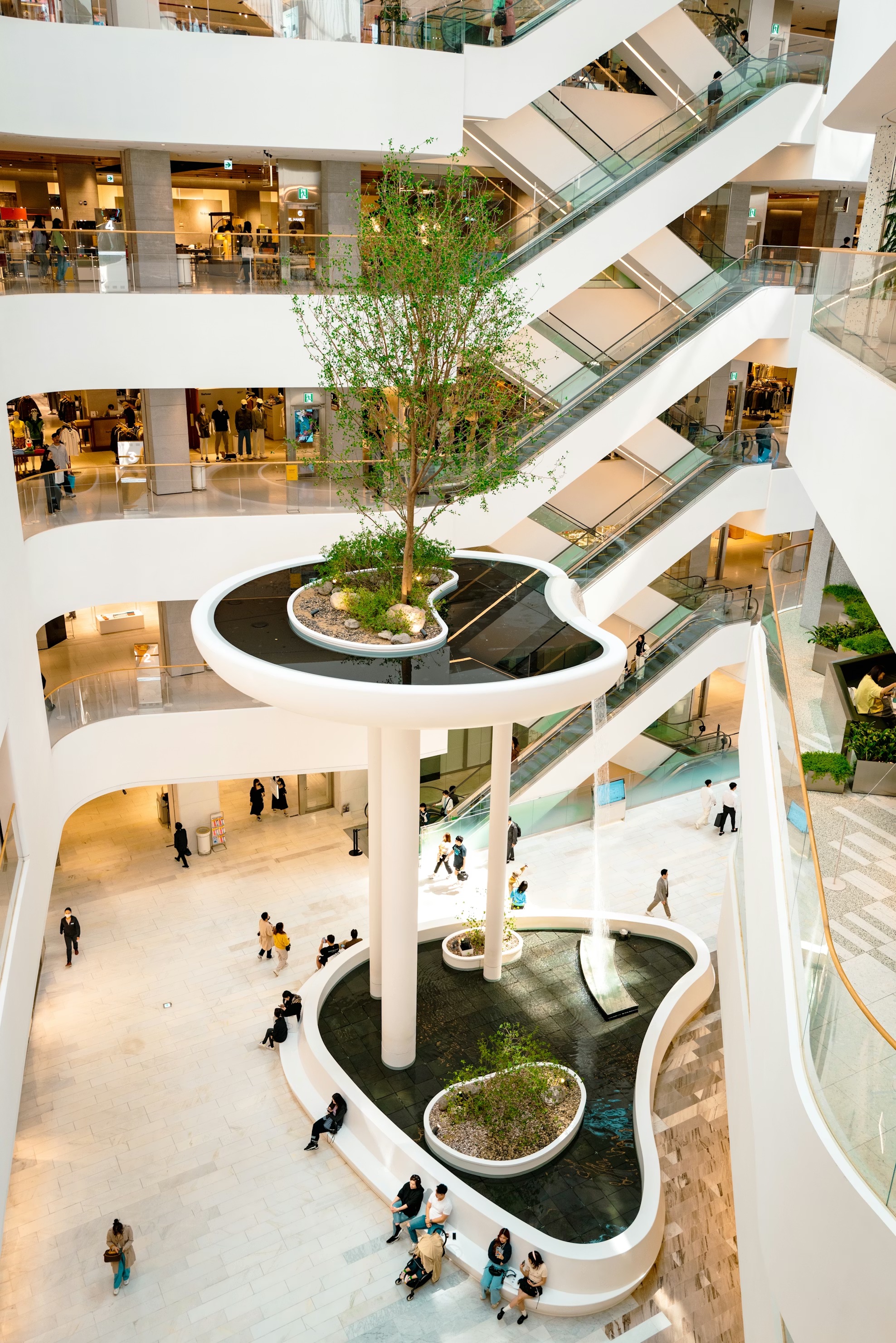 Modern multi-level shopping mall interior with a large indoor planter and water feature surrounded by people sitting and walking near escalators.