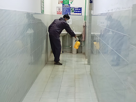 Person wearing protective clothing and mask disinfecting a tiled corridor near a sink with a yellow sprayer.