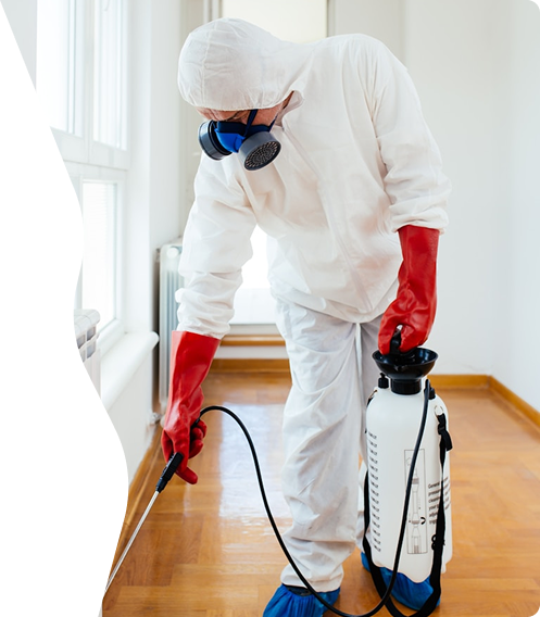 Person in protective suit, red gloves, and respirator spraying disinfectant in a room with wooden floor.