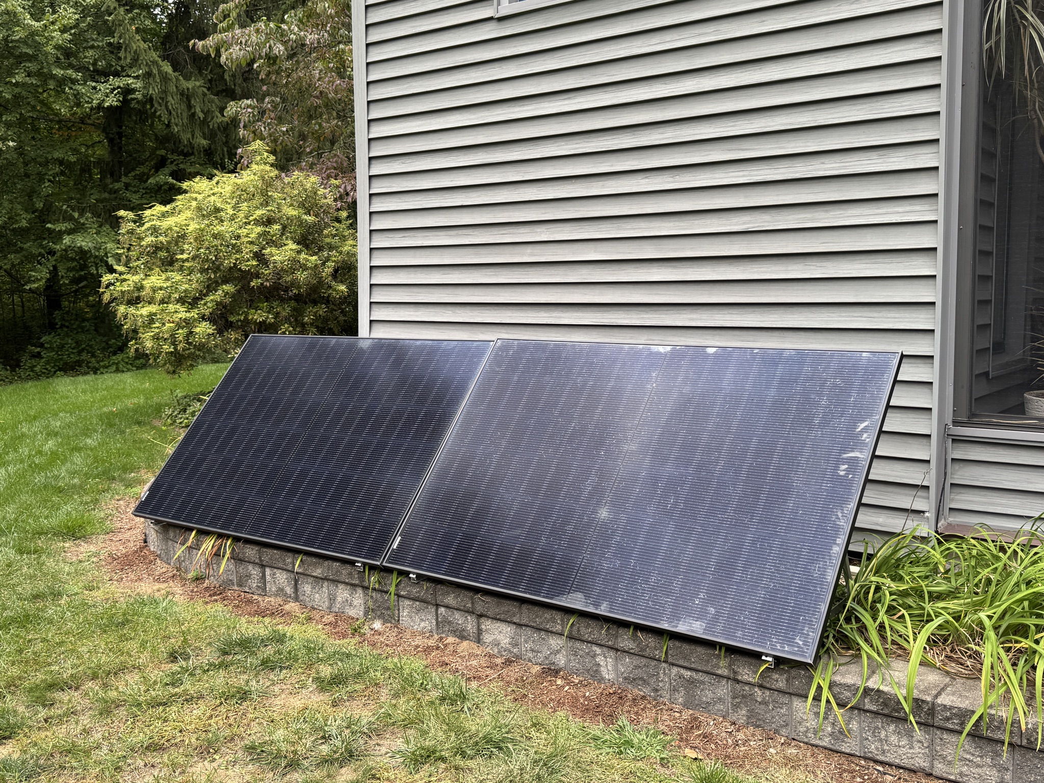 Two black-framed solar panels on a low ground mount beside a gray-sided house