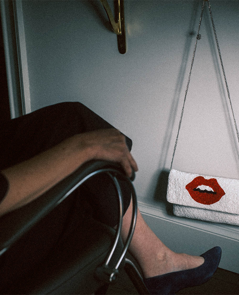 Person sitting on a chair with one leg crossed, wearing blue shoes, next to a wall with a white handbag featuring red lips hanging on a chain.