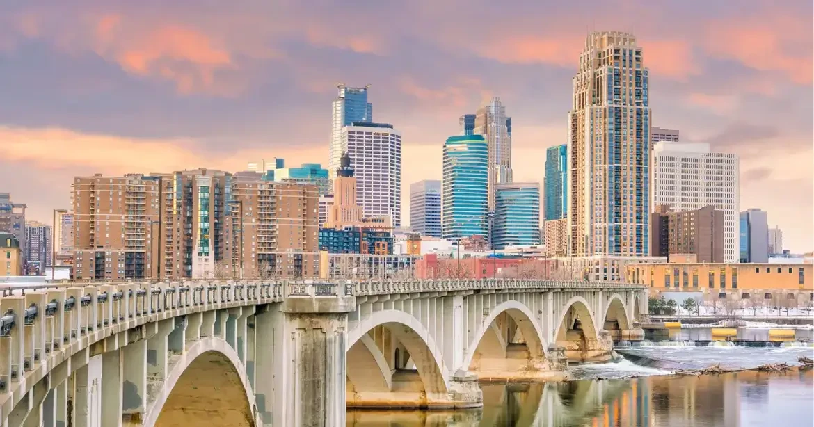 A cityscape of Minneapolis featuring a bridge with multiple arches in the foreground. The skyline includes various modern skyscrapers and buildings under a colorful, cloudy sky.