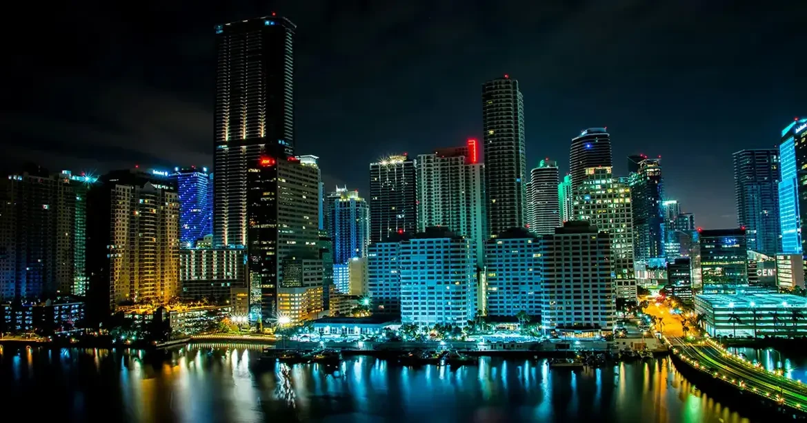 Miami's downtown skyline glowing at night, with illuminated skyscrapers reflected in the water below.