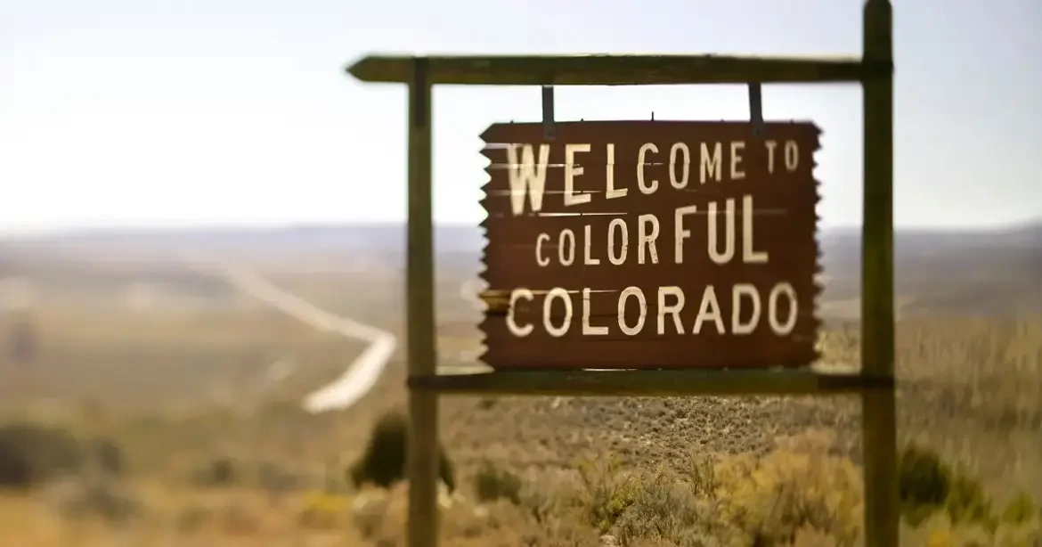 A wooden sign in a rural landscape reads "Welcome to Colorful Colorado" with a blurred background of open fields and a distant road.
