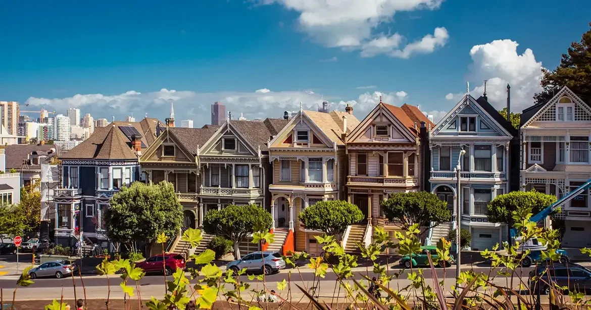 A row of colourful Victorian houses, known as the Painted Ladies, in San Francisco under a clear blue sky, with the city skyline visible behind them and cars parked along the street in front.