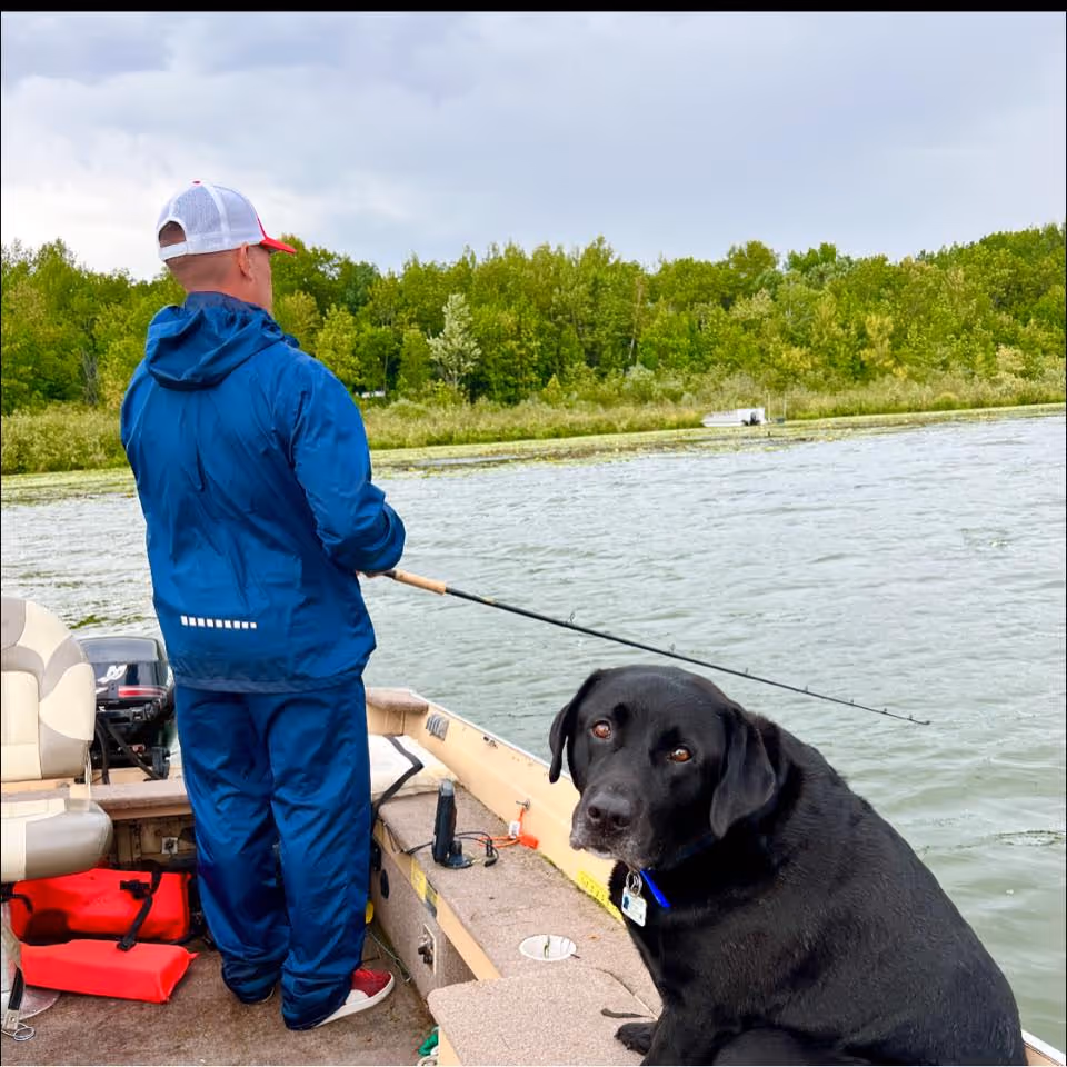 Man in blue rain jacket and cap fishing from a boat with a black Labrador sitting beside him.