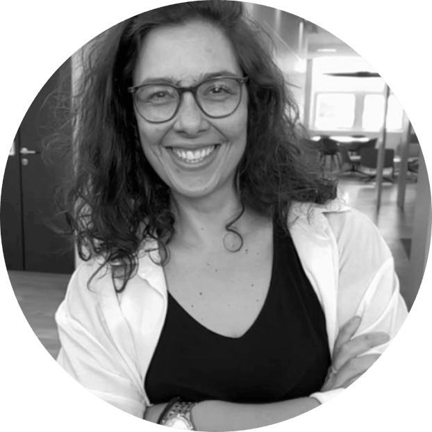 Smiling woman with curly hair, glasses, wearing a black top and white shirt, arms crossed in an office setting.