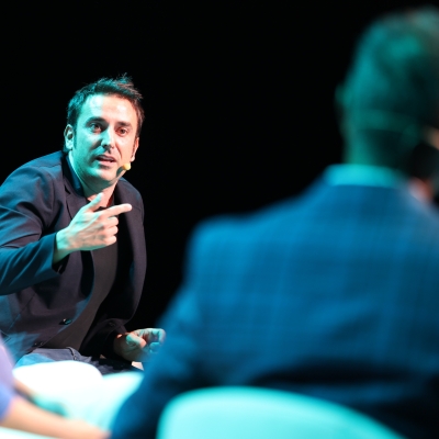 Man in a dark blazer speaking and gesturing during a panel discussion with a blurred man in a checkered jacket in the foreground.