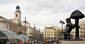 Statue of a bear reaching for a tree in Puerta del Sol with historic buildings and clock tower in the background under a cloudy sky.