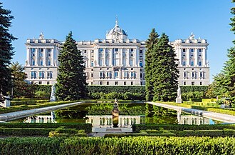 The Royal Palace of Madrid with a large reflecting pond and manicured garden in the foreground under a clear blue sky.