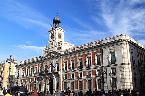 Large historic building with clock tower and flags, crowds gathered in front under blue sky.