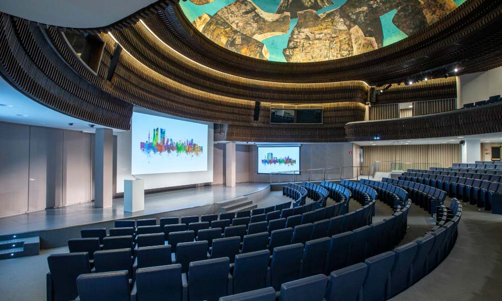 Modern auditorium with curved rows of blue seats, a stage with two large screens displaying a colorful city skyline, and unique ceiling artwork.