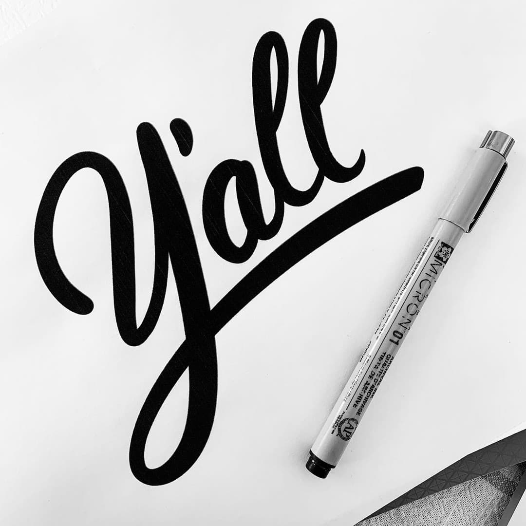Hand-lettered cursive word "Y'all" next to a Micron pen, a hand-lettering sample.