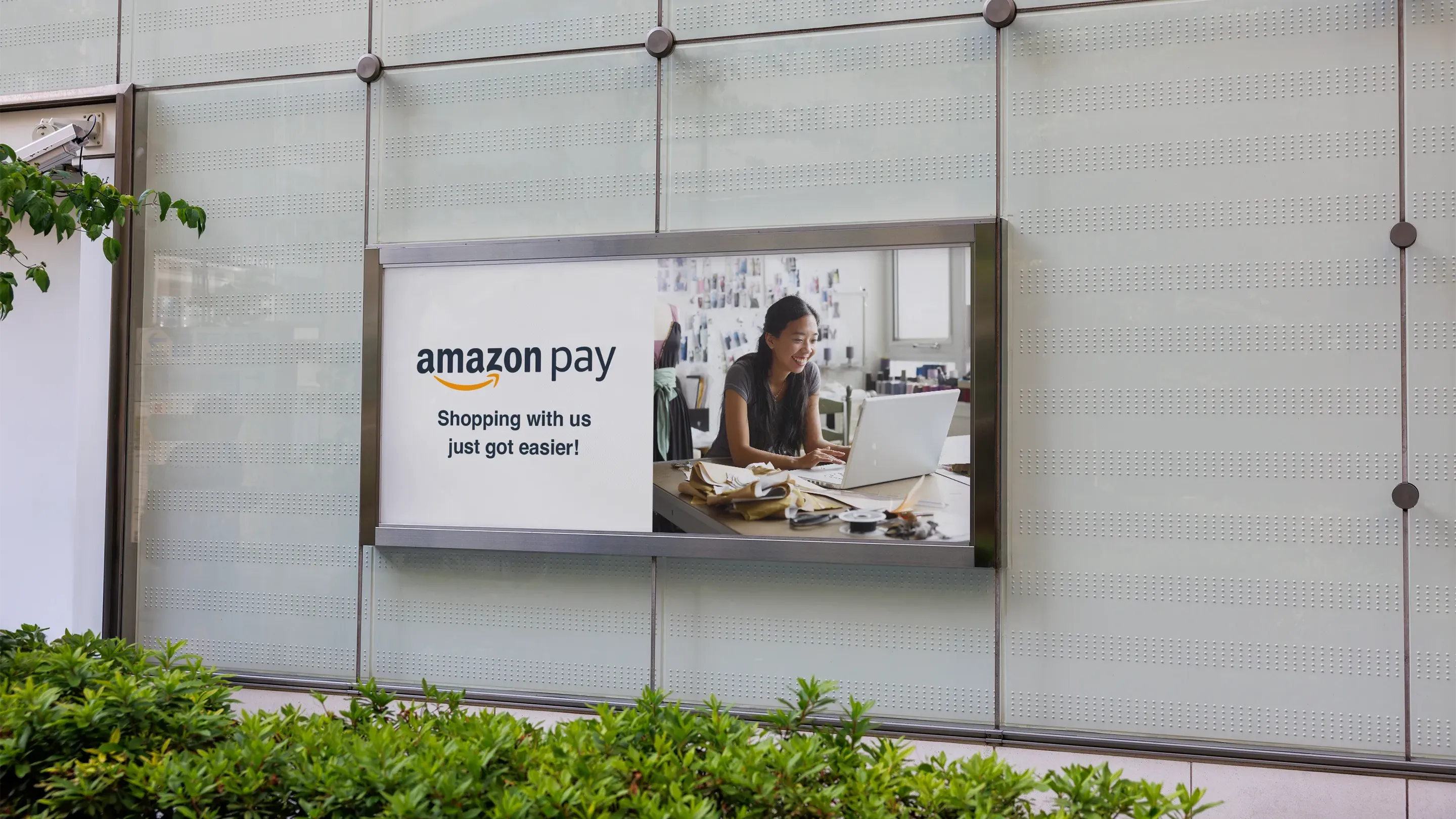 Horizontal Amazon Pay billboard mounted on a glass exterior wall, featuring a small-business owner working on her laptop at a sewing studio bench beside the "Amazon Pay — Shopping with us just got easier!" headline.