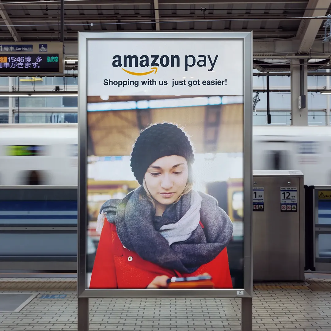 Vertical Amazon Pay out-of-home placement at a Japanese train platform, featuring a woman in a red coat checking her phone alongside the "Amazon Pay — Shopping with us just got easier!" headline, with bullet trains visible in motion behind it.