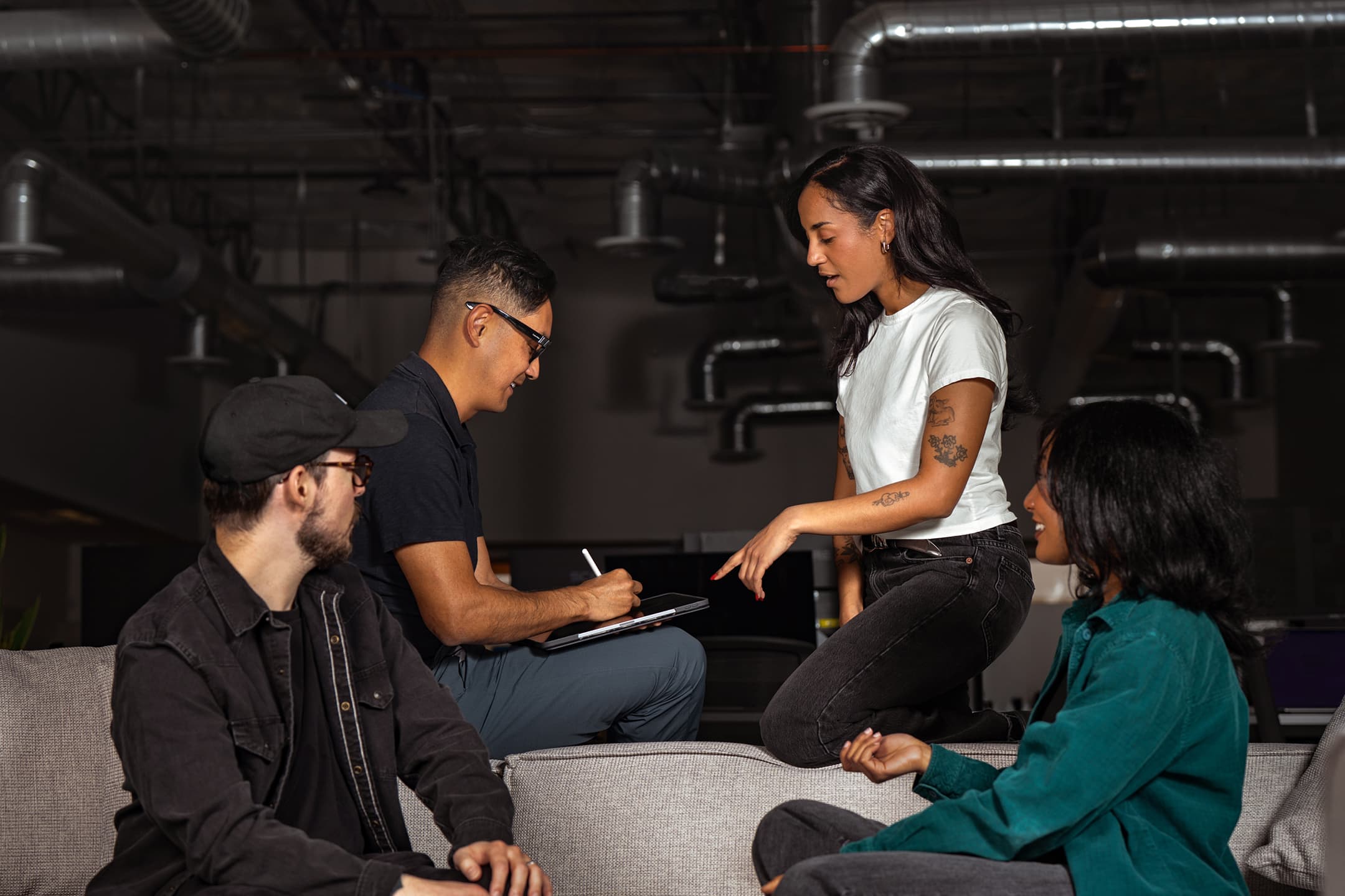 Three creative professionals in a working session on a couch — one taking notes on a tablet while another presents an idea, in a dimly lit modern office.