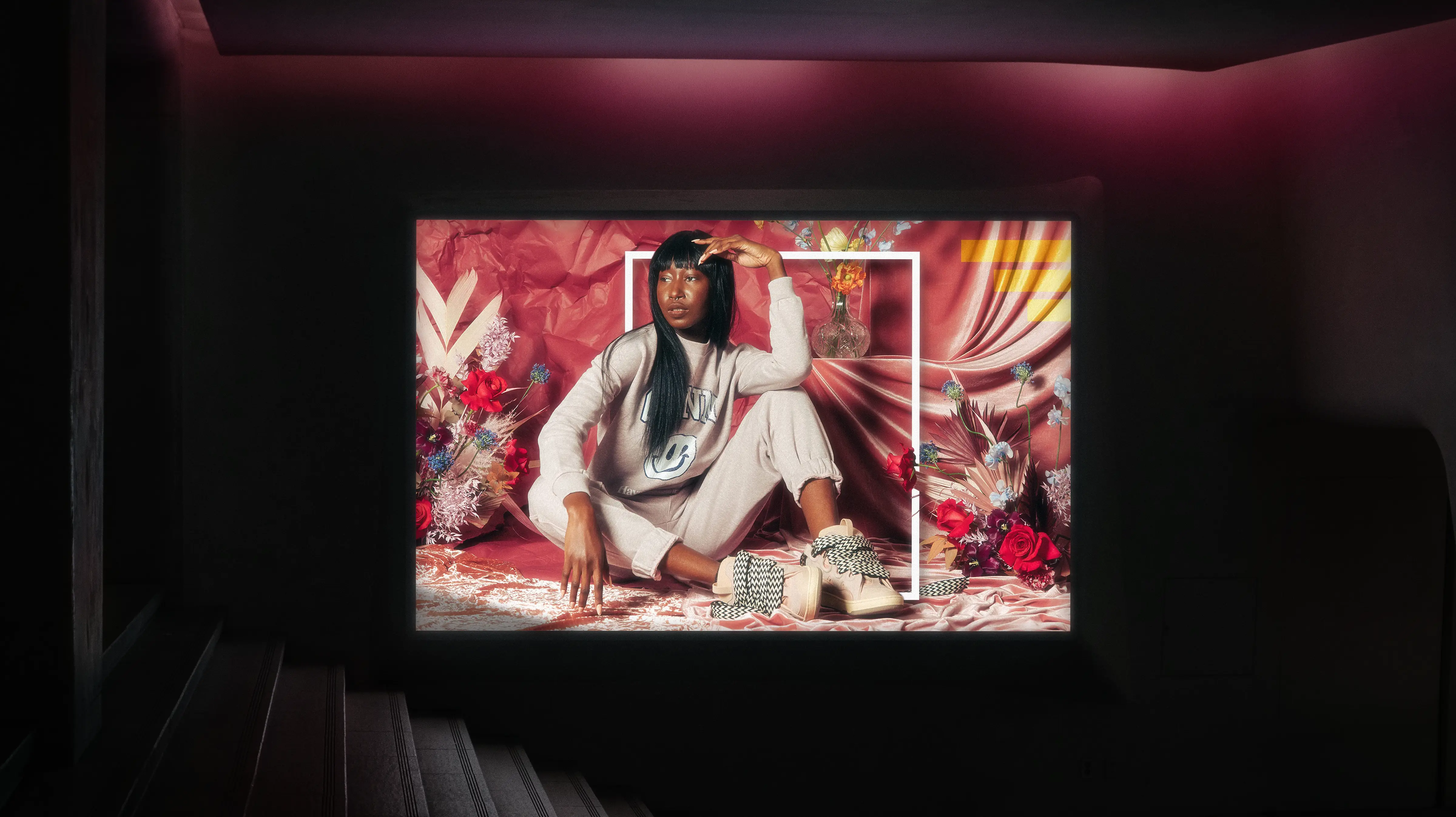 VRSNL campaign image art designed by Alex Mera — a model in a cream graphic sweatsuit and checkered sneakers, seated against a red draped-fabric backdrop with florals and a bold white frame overlay, shown projected in a screening room.
