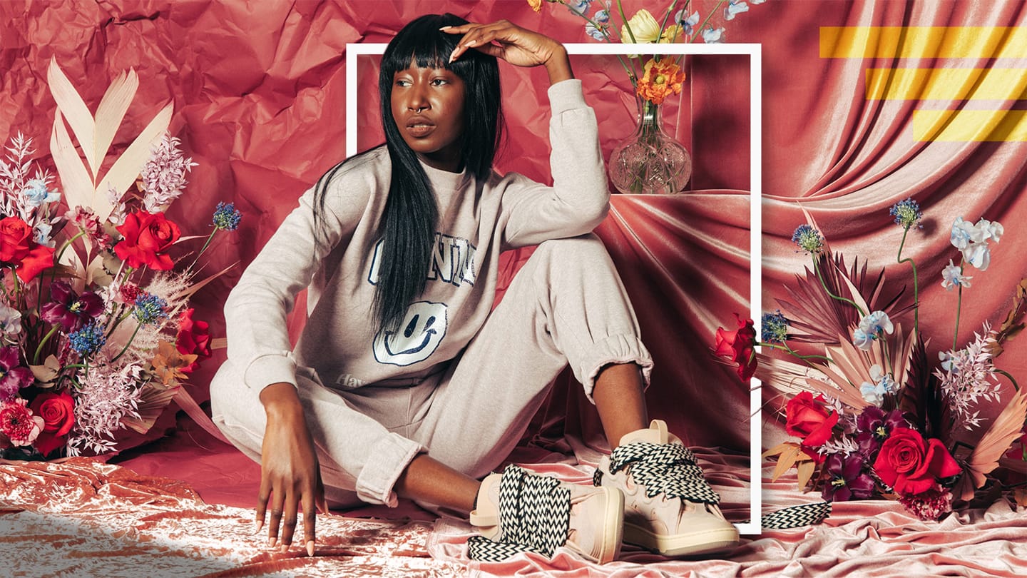 VRSNL editorial image art directed by Alex Mera — a model in a cream graphic sweatsuit and checkered sneakers, seated against a red draped-fabric backdrop with florals and a bold white frame overlay.