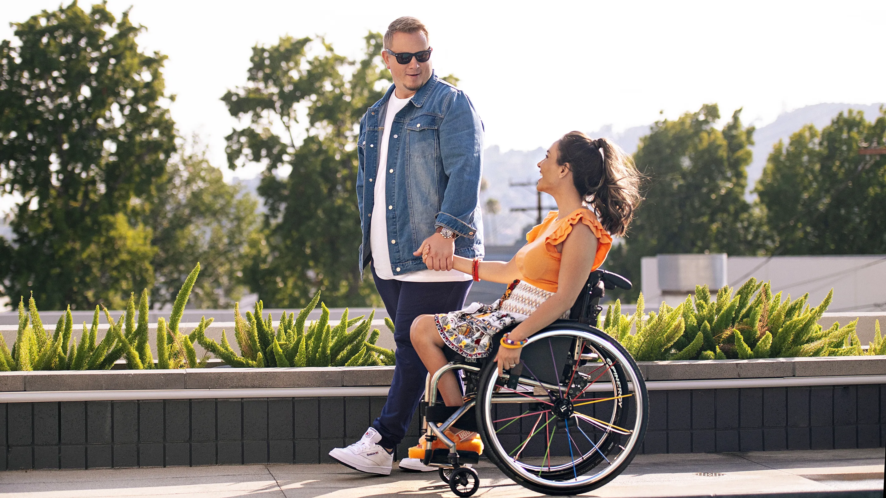 Man and woman holding hands and smiling on a rooftop patio; she wears an orange top and sits in a wheelchair.