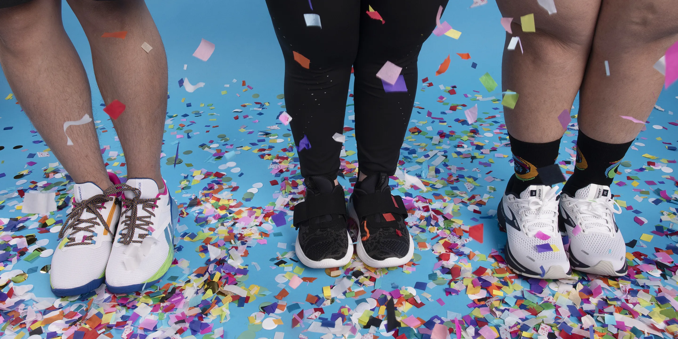 Close-up of three pairs of legs and sneakers standing on a blue floor covered in colorful confetti, showcasing Reebok, ASICS, and Brooks running shoe styles from the Zappos x Special Olympics collection.