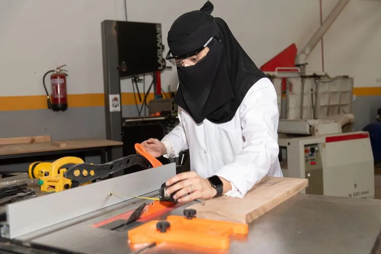 Woman wearing a niqab and safety glasses operating a table saw in a workshop.