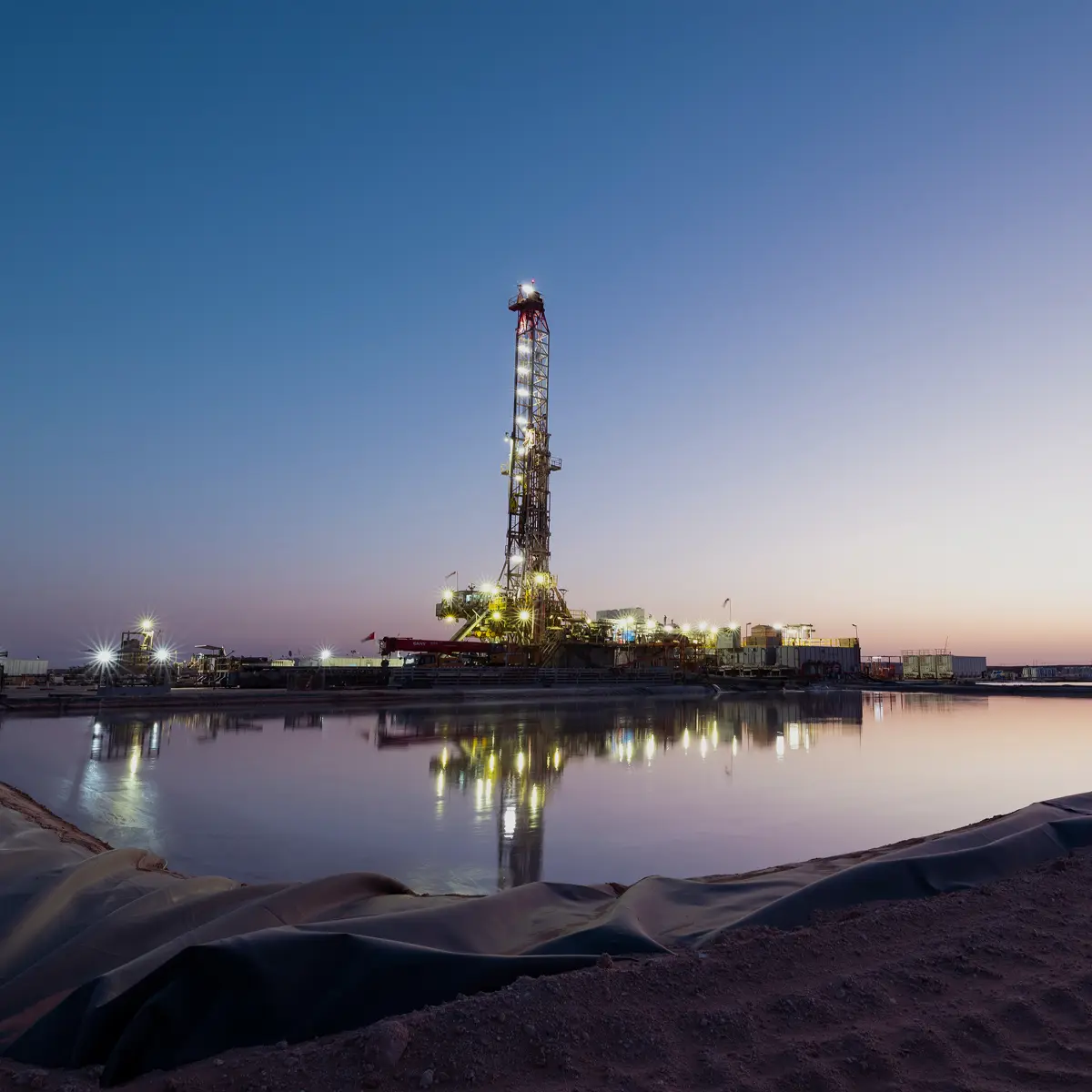 Illuminated drilling rig reflected in water at dusk