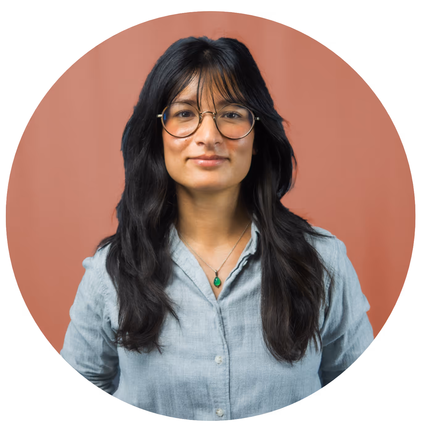 Young woman with long dark hair, round glasses, and a light blue buttoned shirt, standing against a muted reddish background.