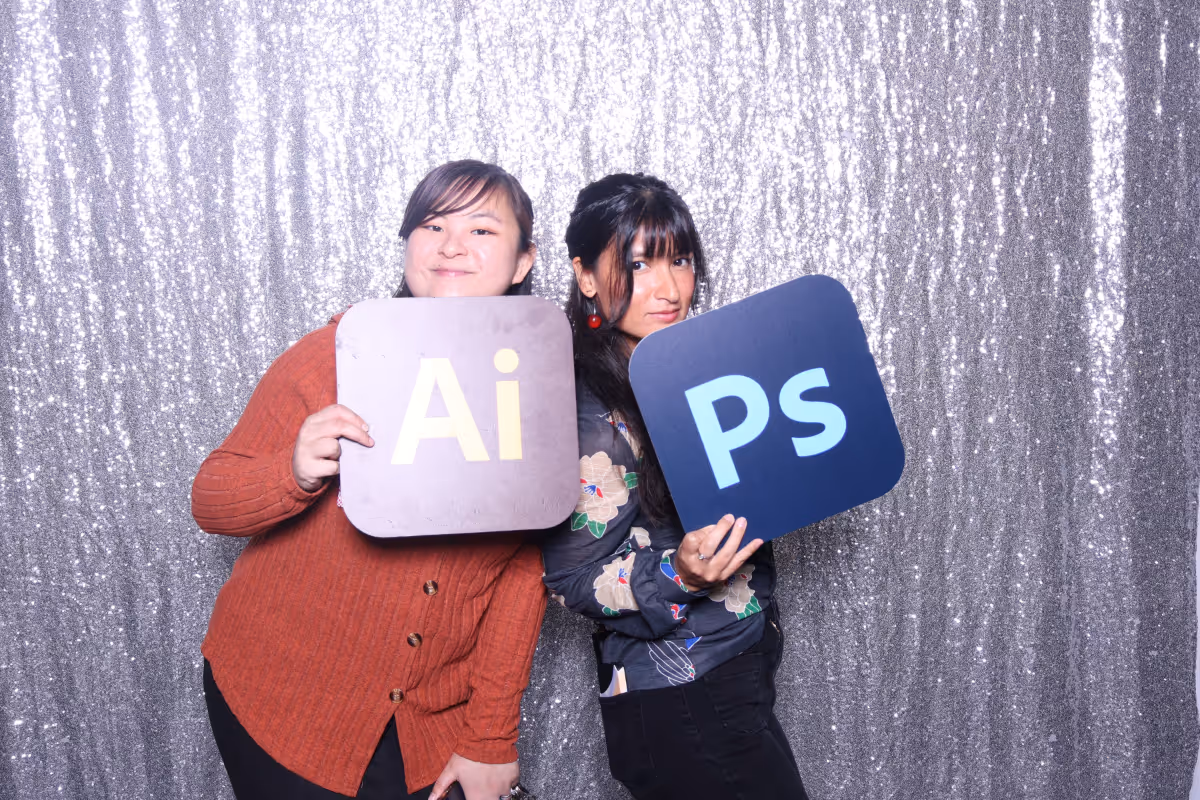 Two women standing back to back holding Adobe Illustrator and Photoshop logo signs against a sparkly silver backdrop.