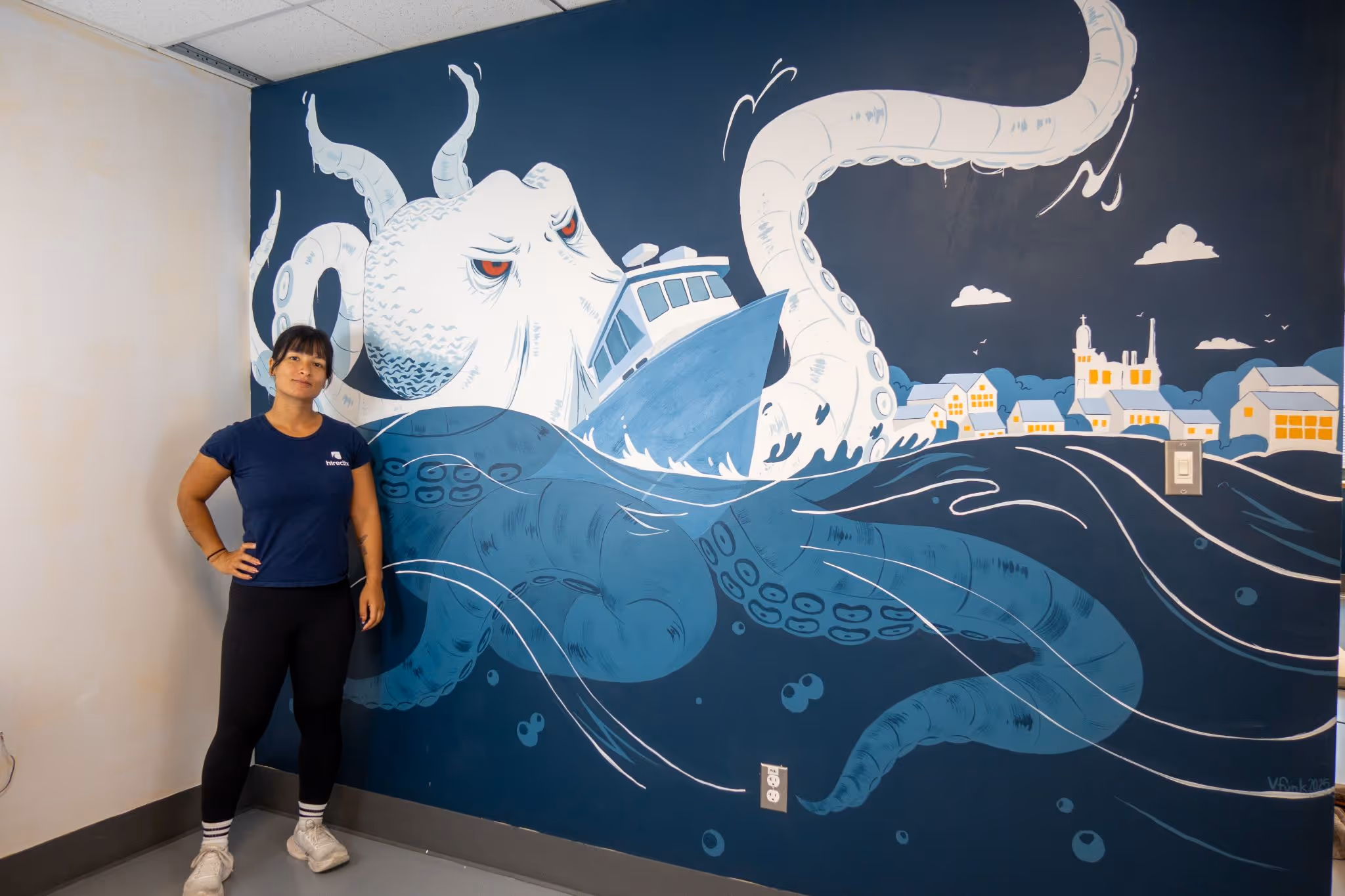 Woman standing beside a mural of a giant octopus attacking a boat near a seaside village at night.