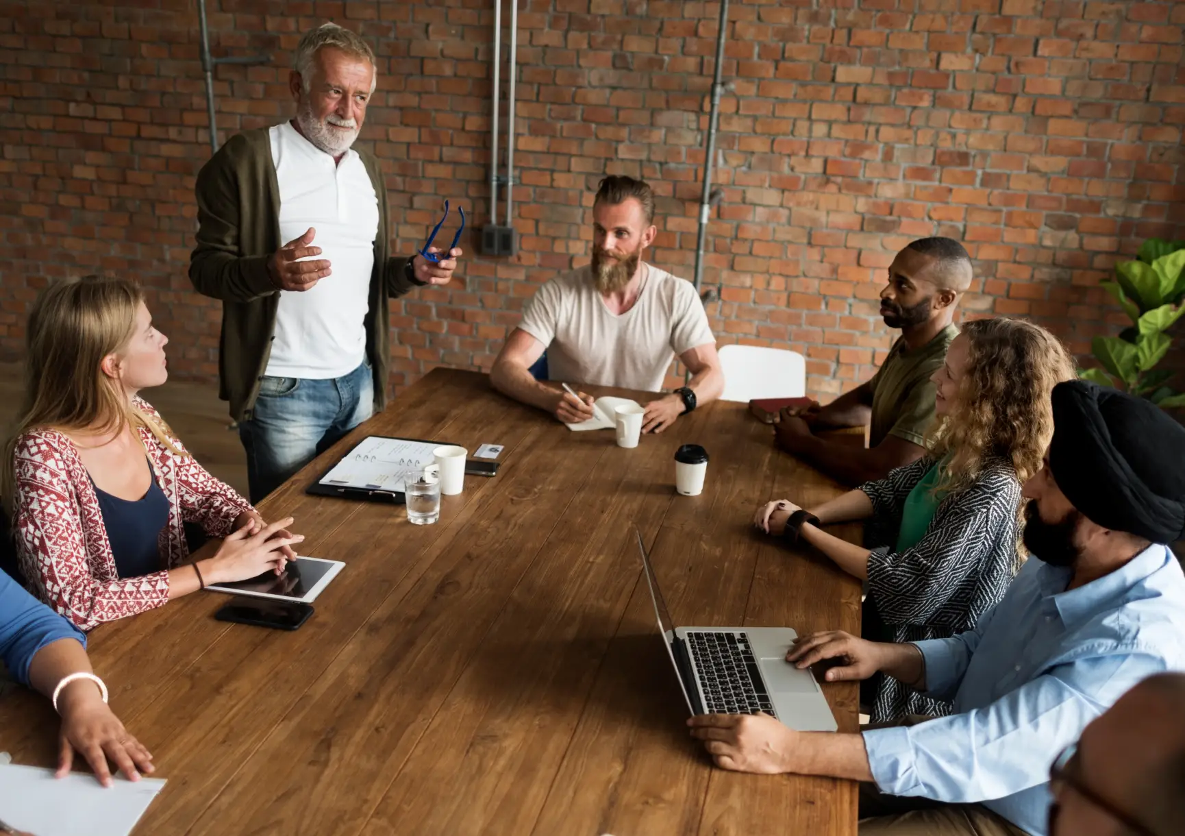 A diverse group of people seated around a wooden table listens as an older man standing speaks, holding glasses in one hand.