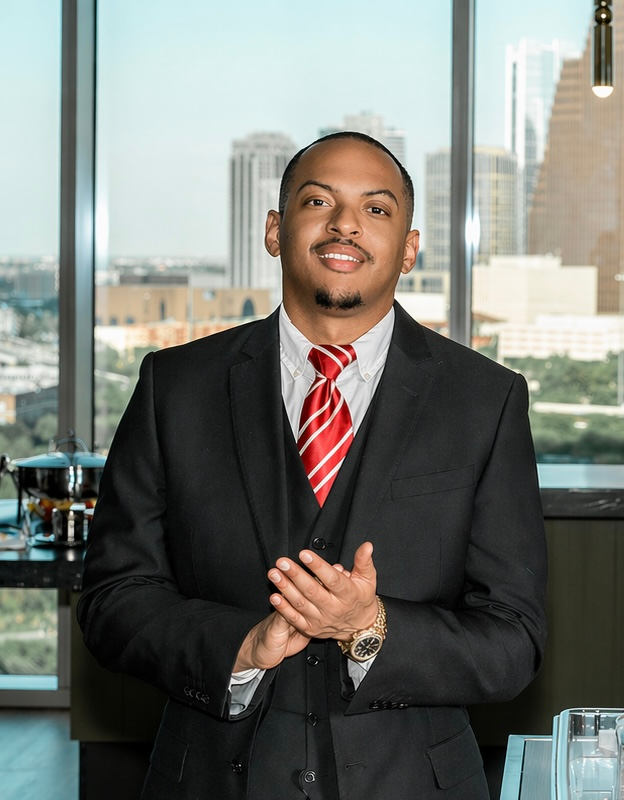 Man in black suit, white shirt, and red striped tie standing indoors with city buildings visible through large windows behind him.