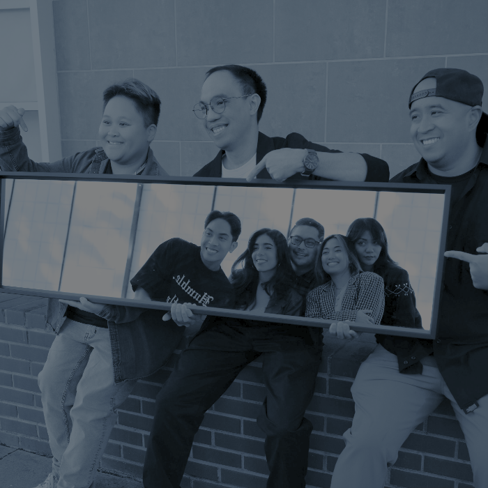 Three people smiling and holding a large mirror reflecting five people posing together in front of a tiled wall.