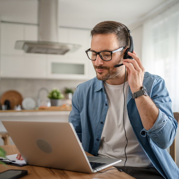 Man wearing headset and glasses sits at table using laptop for a call in a bright kitchen.
