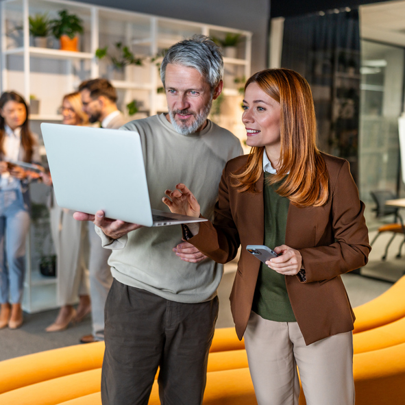 Man and woman looking at a laptop together in a modern office with colleagues in the background.