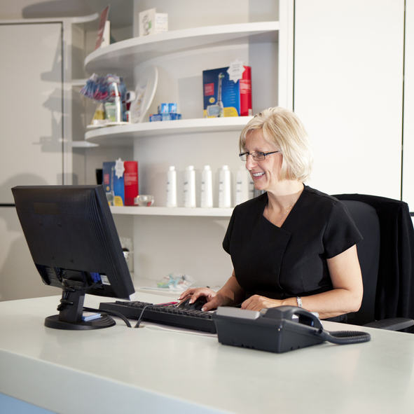 Smiling woman with glasses working on a computer at a desk in a modern office or clinic.