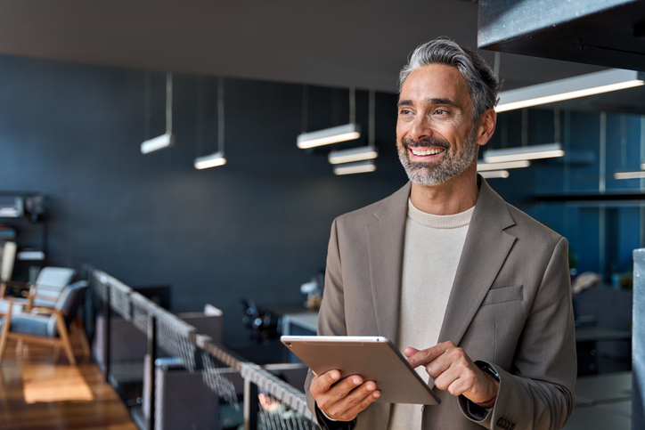 Smiling man in beige blazer holding tablet in modern office with dark blue walls and hanging lights.