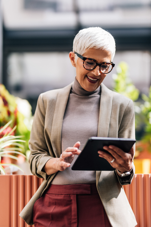 Smiling woman with short white hair using a tablet while leaning on a ledge in a bright indoor space.
