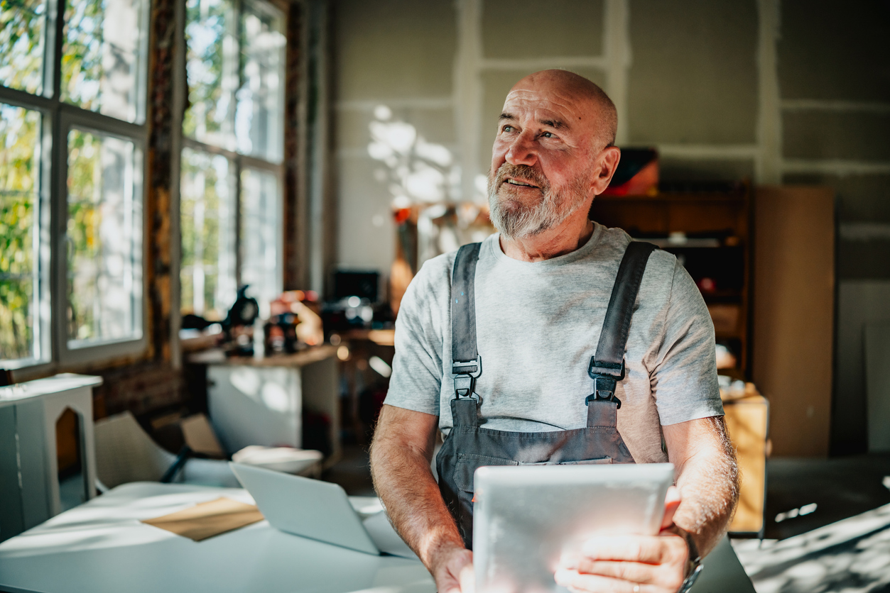 Smiling elderly man in overalls holding a tablet in a bright, modern workshop space.