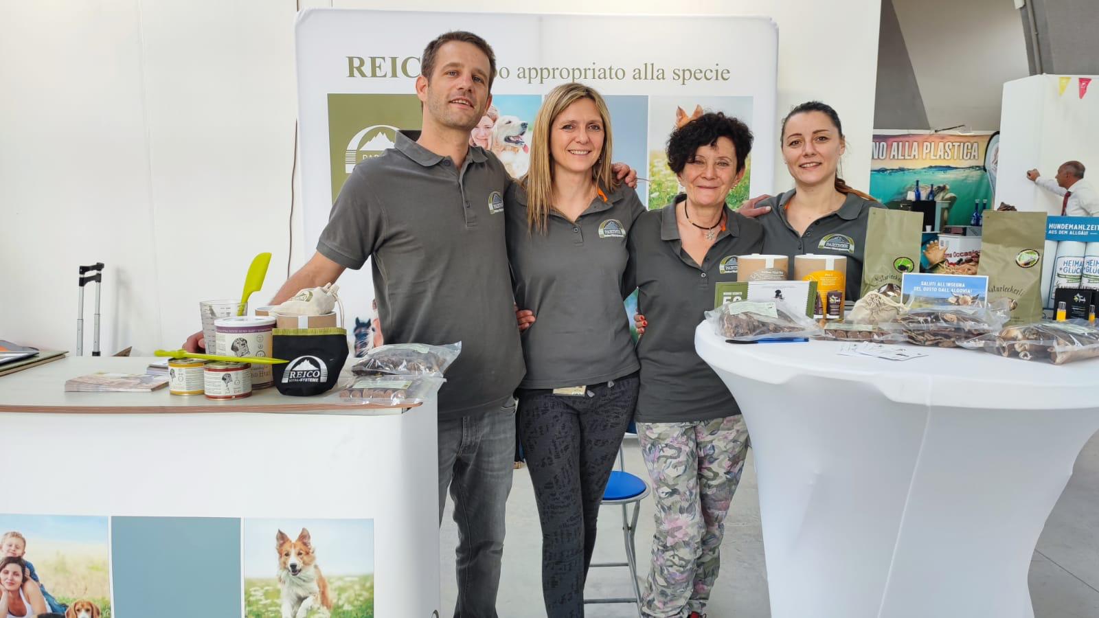 Four people wearing matching grey polo shirts standing behind two tables displaying pet food products and promotional materials.