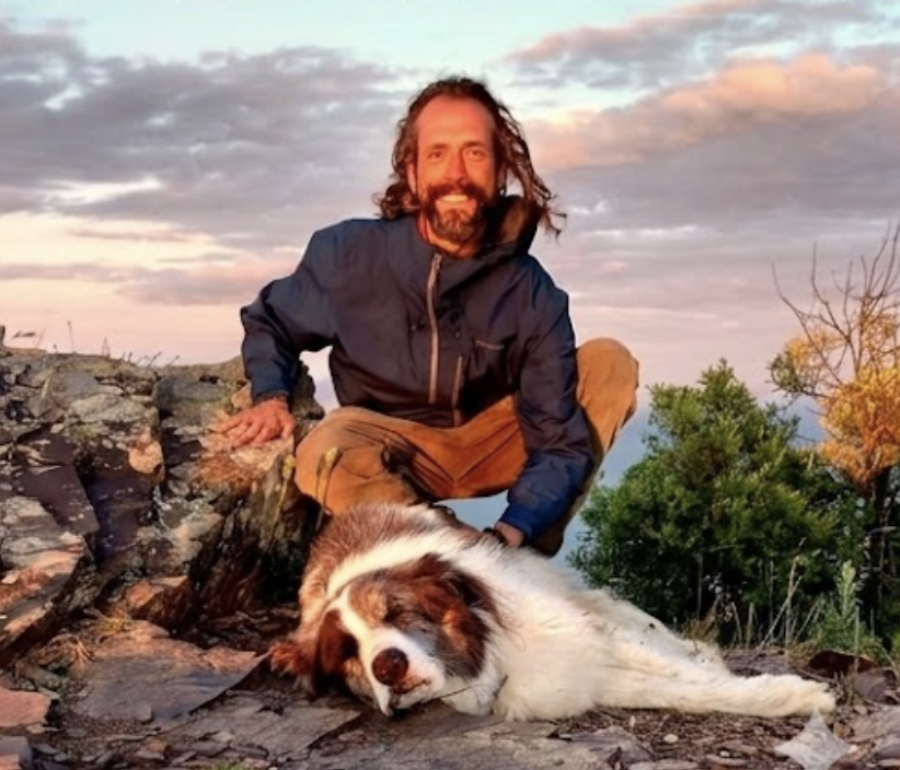 Man in a blue jacket squatting on rocky ground next to a large white and brown dog lying down with eyes closed during a sunset.